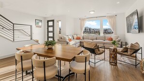 A modern living room with a wooden dining table, beige chairs, a sofa set, and a large window with a city view. - Always An Adventure 4 Bedroom Townhouse (St. George)