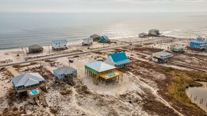 Una zona costera con varias casas de playa sobre pilotes, un edificio principal con techo azul y una playa de arena con una carretera cerca.