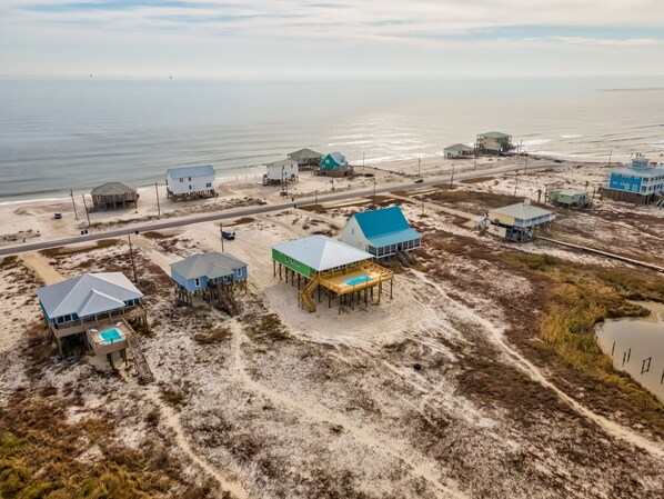 Una zona costera con varias casas de playa sobre pilotes, un edificio principal con techo azul y una playa de arena con una carretera cerca.