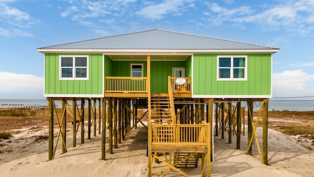 Una casa verde de madera con balcón y una escalera sobre pilares, ubicada en una playa arenosa con vista al océano.