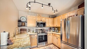 A modern kitchen with wooden cabinets, stainless steel appliances, a granite countertop, and a patterned rug. - 5959 Hidden River Lodge 2 Bedroom Condo (Keystone)