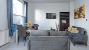 A modern living room with a grey sofa, a dining table with chairs, a TV mounted on the wall, and a bookshelf.