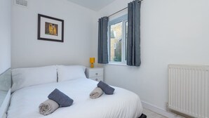 A neatly made bed with rolled towels, a framed picture on the wall, a bedside table with a lamp, and a window with curtains.