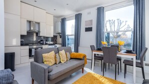 A modern living room with a grey sofa, a dining table with chairs, a kitchen area, and a large window with curtains.