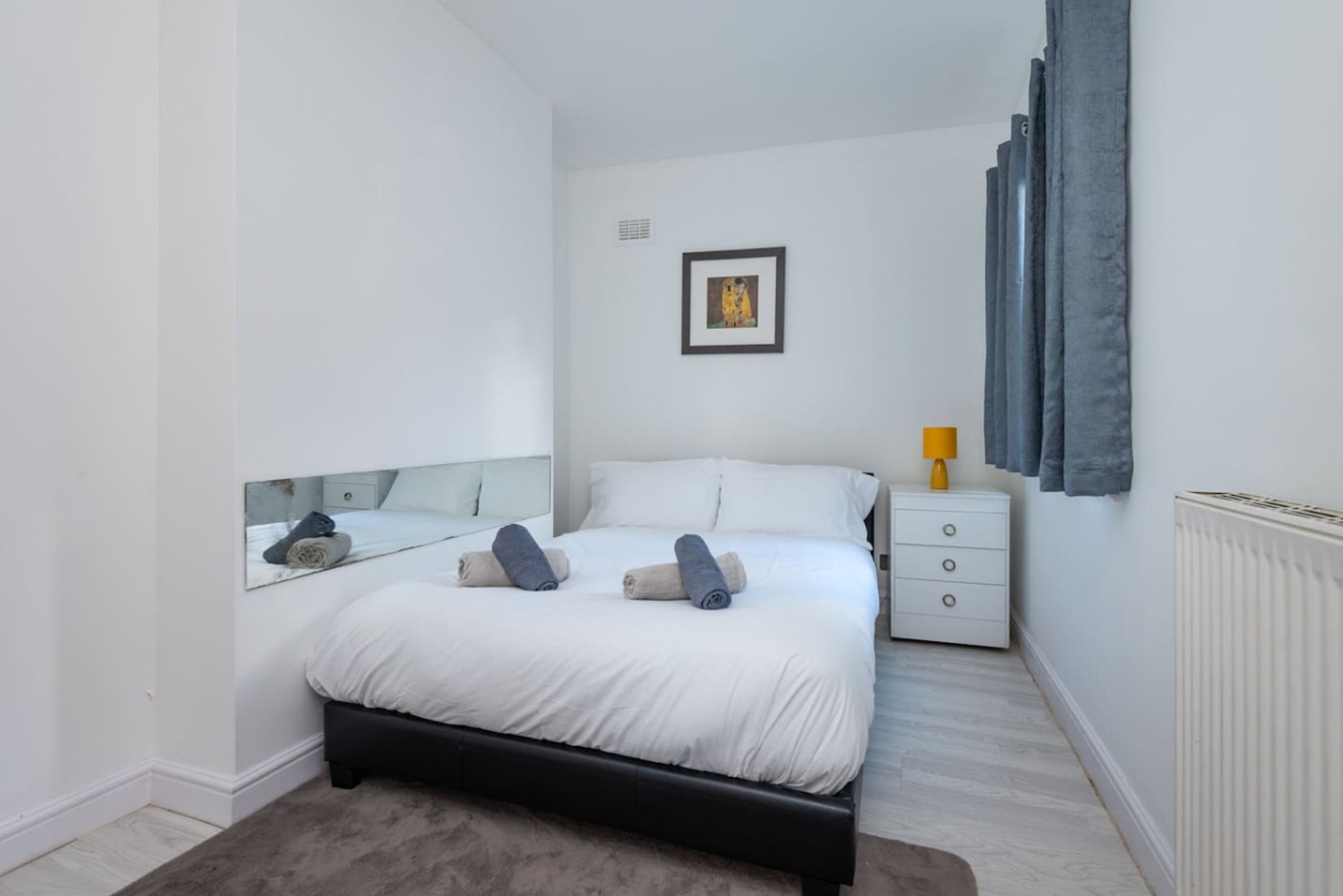 A bedroom with a bed, a bedside table, a lamp, a mirror, and a framed picture on the wall.