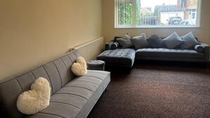 A living room with a grey sofa, a smaller grey loveseat, and a round ottoman. There is a large window with a view of buildings and cars outside. - 3 Bed House - Sleeps 6 - Parking - Wifi (Leicester)