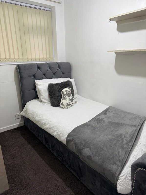 A small, neatly arranged bedroom with a grey upholstered headboard, a white textured bedspread, and a grey throw blanket. There is a white pillow with a black and white design, and a fluffy grey pillow. A small shelf is mounted on the wall above the bed.