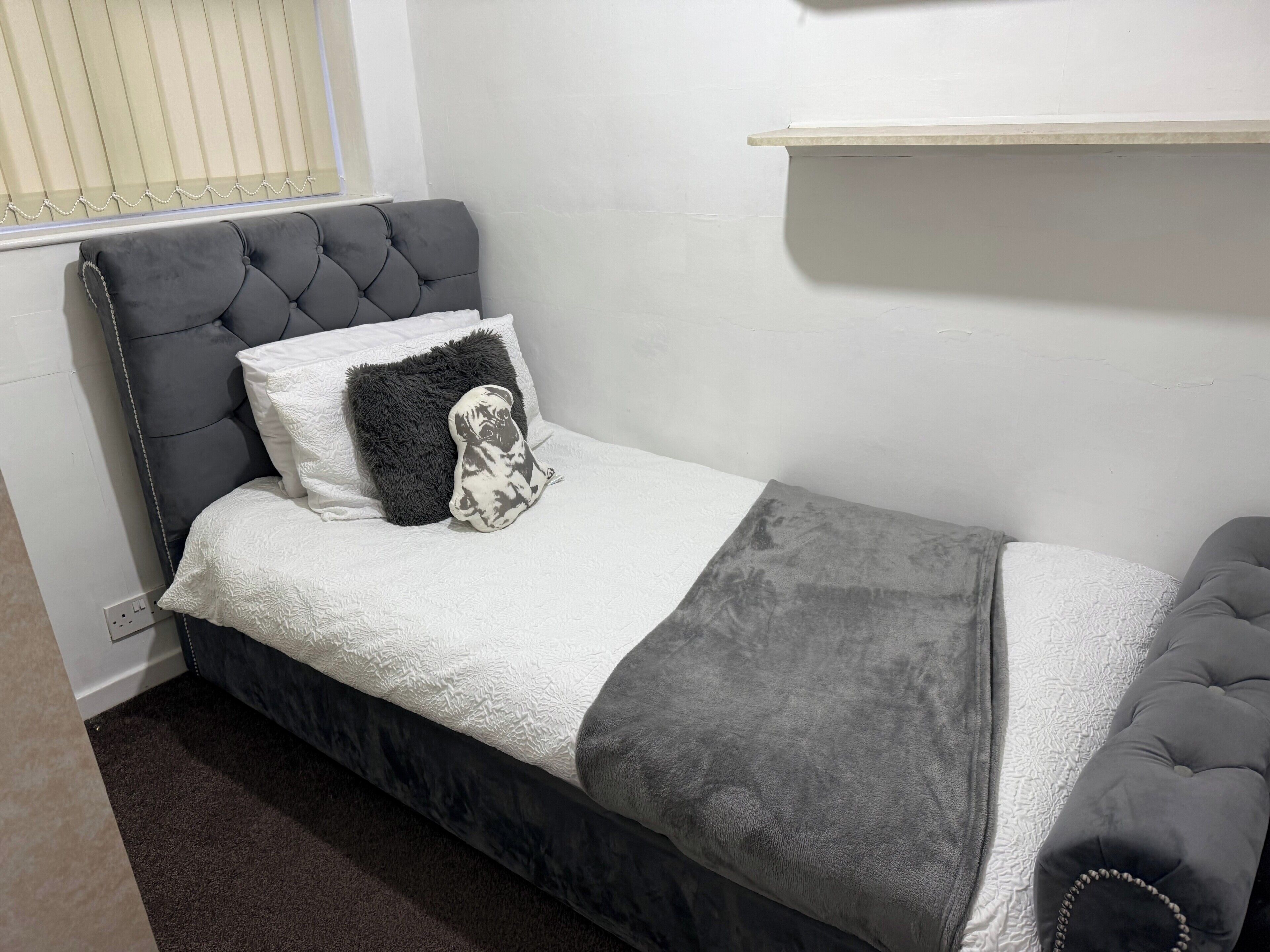 A small, neatly made bed with a grey upholstered headboard, white and grey bedding, and a decorative pillow.