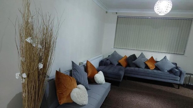 A modern living room with a grey sectional sofa, multiple cushions, and a decorative vase with dried branches. - 3 Bed House - Sleeps 6 - Parking - Wifi (Leicester)