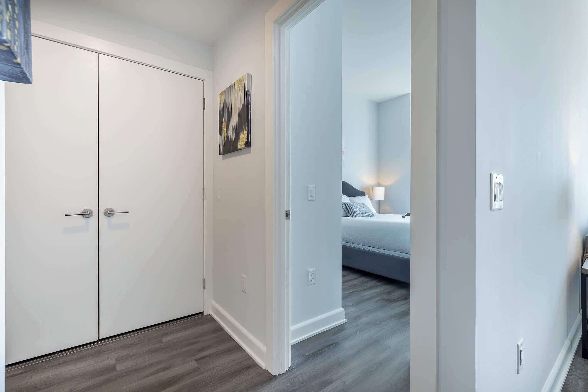 A hallway leading to a bedroom with a bed, a nightstand, and a lamp. There is a door on the left and a painting on the wall.