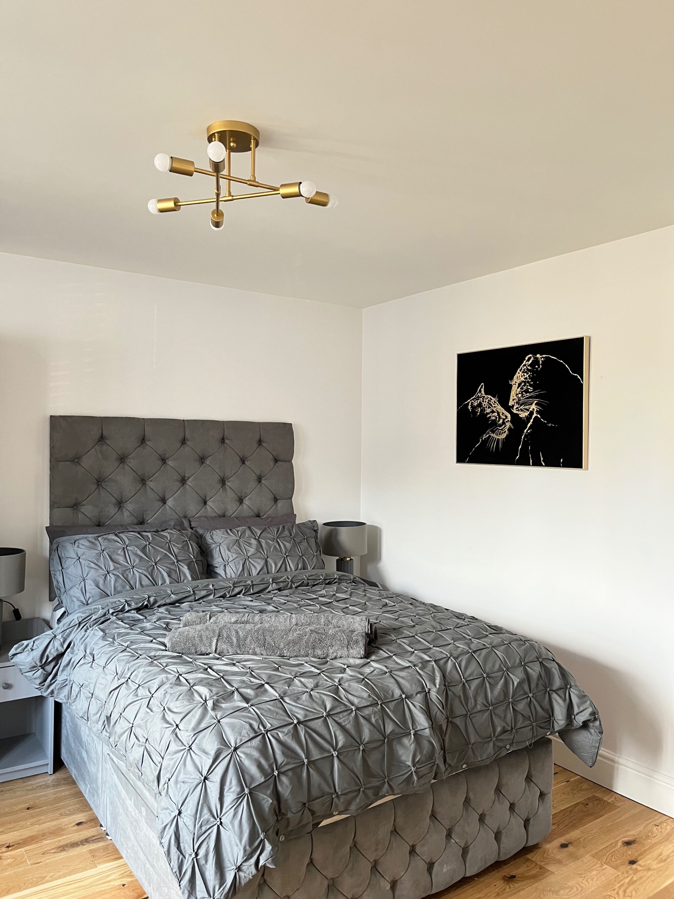 A bedroom with a tufted headboard, a grey quilted bedspread, a modern chandelier, and a framed artwork on the wall.