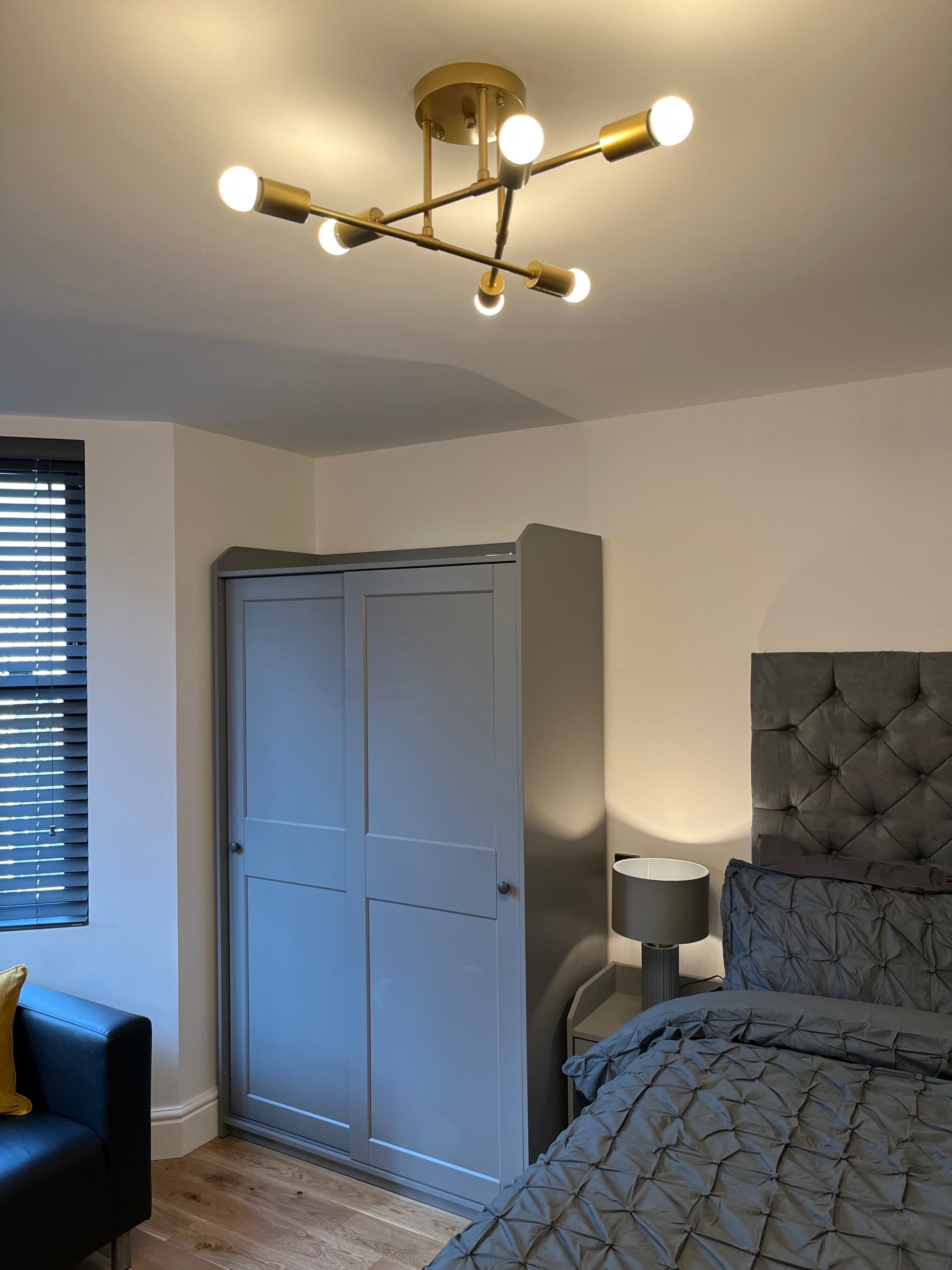 Ein Schlafzimmer mit einem grauen Kleiderschrank, einem Bett mit grauem Kopfteil, einem Nachttisch mit Lampe und einer Deckenleuchte.