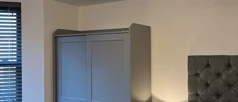 Ein Schlafzimmer mit einem grauen Kleiderschrank, einem Bett mit grauem Kopfteil, einem Nachttisch mit Lampe und einer Deckenleuchte.