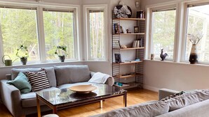 A living room with a grey sofa, a coffee table, and a bookshelf. There are large windows with plants on the windowsill.