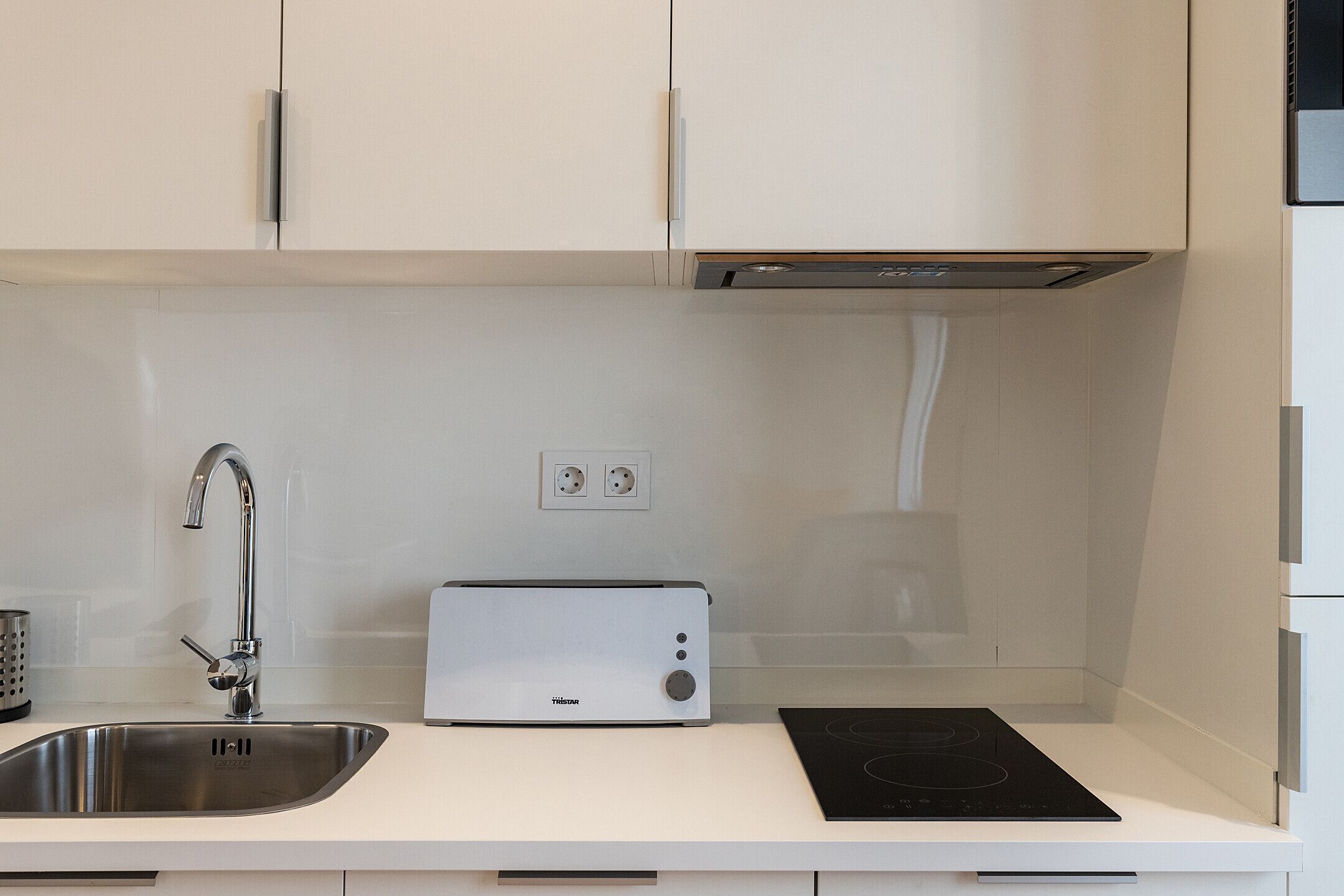 Una cocina moderna con gabinetes blancos, un lavamanos de acero inoxidable, un tostador y una placa de cocción por inducción negra.