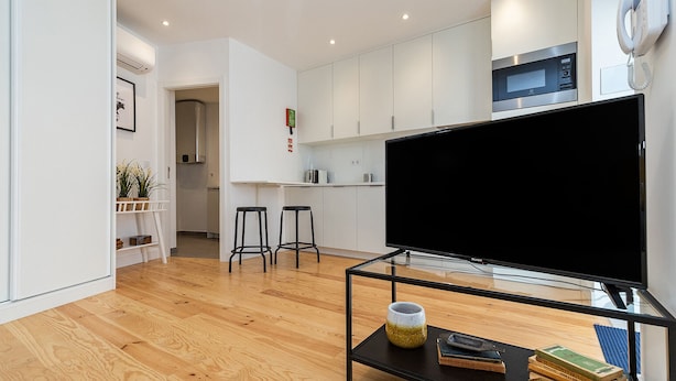 A modern living room with a large flat-screen TV, a coffee table with books and a vase, and a kitchen area with white cabinets and a microwave. - Belem Design Studio 2 by Homing (Lisbon)