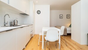 A modern kitchen with white cabinets, a stainless steel sink, and a kettle. There is a dining area with a white table and two chairs. A bed with a green pillow is visible in the background. - Belem Design Studio 1 by Homing (Lisbon)
