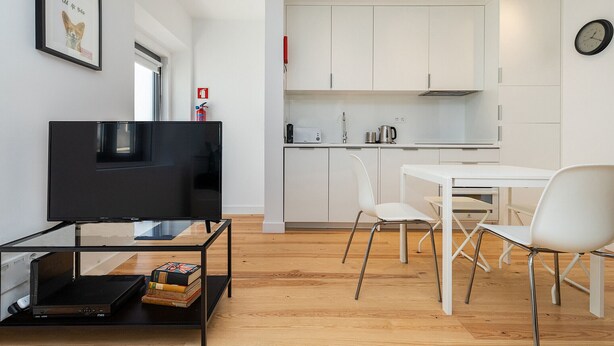 A modern living room with a flat-screen TV, a coffee table, and a bookshelf. There is a dining area with a white table and chairs. The kitchen is visible in the background with white cabinets and a clock on the wall. - Belem Design Studio 1 by Homing (Lisbon)