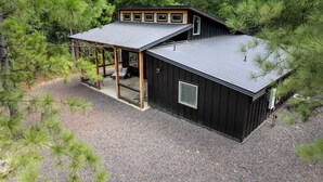 Eine Holzhütte mit überdachter Veranda, einer kleinen Terrasse und einem Schotterweg, umgeben von Kiefern.
