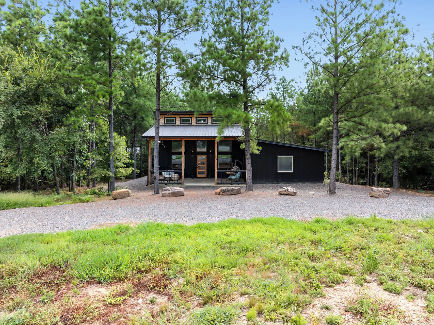 A modern cabin with a large front porch, surrounded by a gravel driveway and a grassy area.