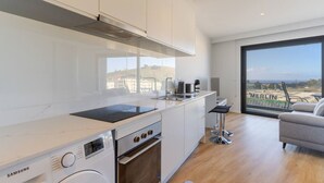 Una cocina moderna con armarios blancos, una campana extractora de acero inoxidable, un horno empotrado y una lavadora Samsung. Hay un taburete de bar y vistas de un paisaje urbano a través de amplias ventanas.