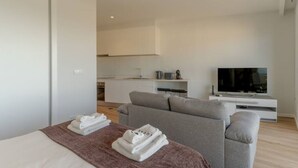 Una habitación de hotel moderna con cama, sofá gris, televisión sobre un mueble blanco y una kitchenette al fondo.