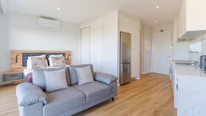 Una sala moderna con un sofá gris, una cama con cabecera de madera y una cocina con refrigerador de acero inoxidable.