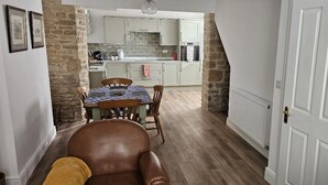 A cozy room with a brown leather chair, a dining table with chairs, a kitchen area with white cabinets, and a stone wall. - Stylish 2-bed House in Stamford Town Centre (Stamford)