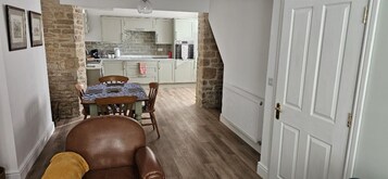 Una habitación acogedora con un sillón de cuero marrón, una mesa de comedor con sillas, una zona de cocina con armarios blancos y una pared de piedra.