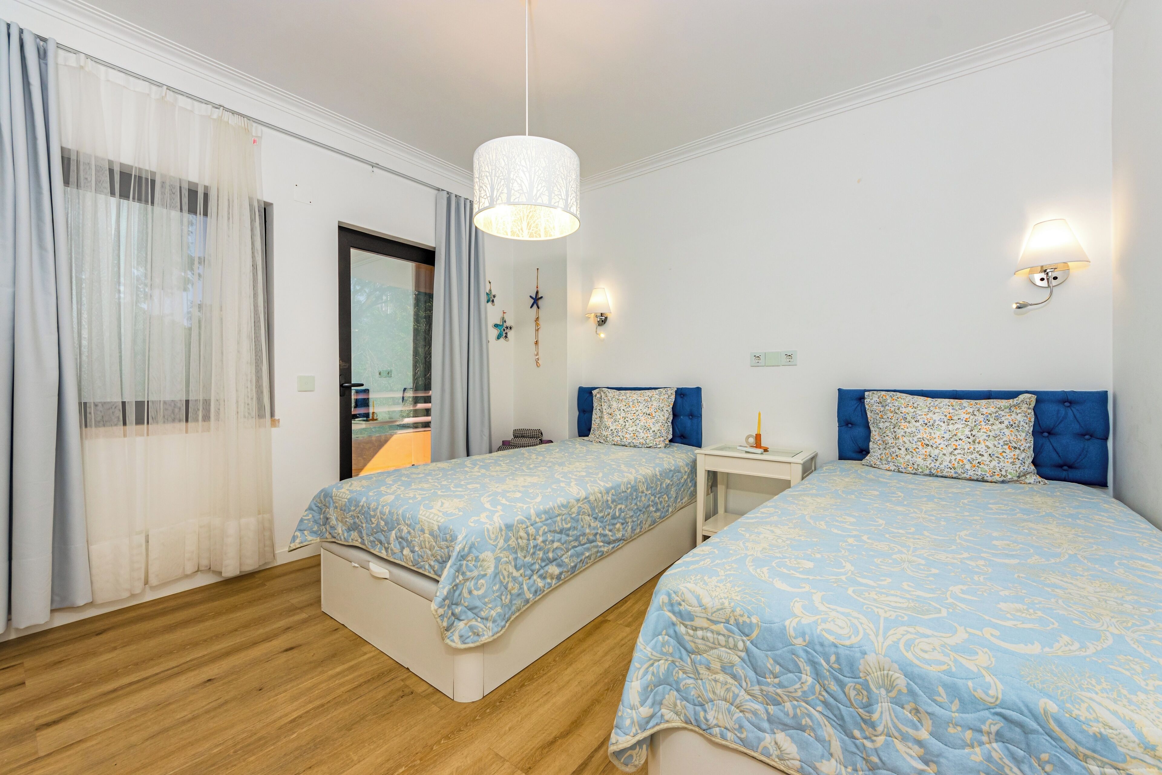 A hotel room with two beds, a wooden floor, a large window with sheer curtains, a white ceiling, and a hanging light fixture.