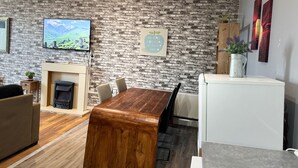 A modern living room with a brick wall, a flat-screen TV, a fireplace, a wooden dining table, and a white refrigerator. - Citycenterapt-4br-wifi-freeparking-retroarcade (Newcastle-upon-Tyne)