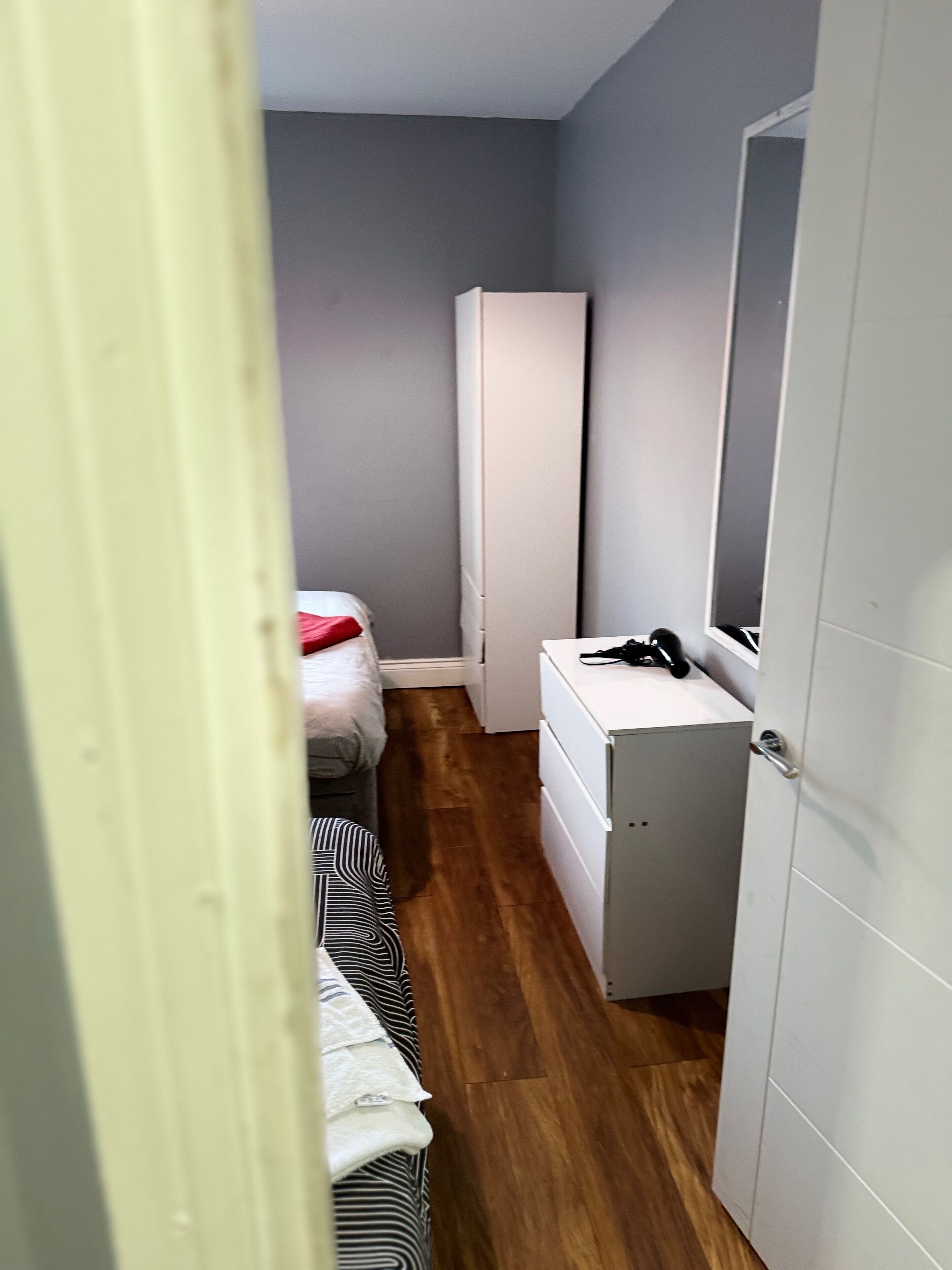 A narrow room with a bed, a wardrobe, and a desk with a mirror.