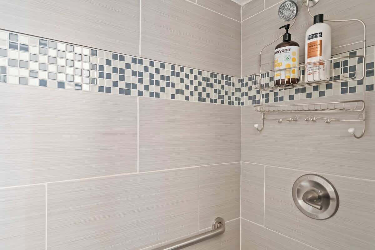 Área de chuveiro com parede revestida em azulejo, barra de apoio e prateleira com produtos de higiene pessoal.
