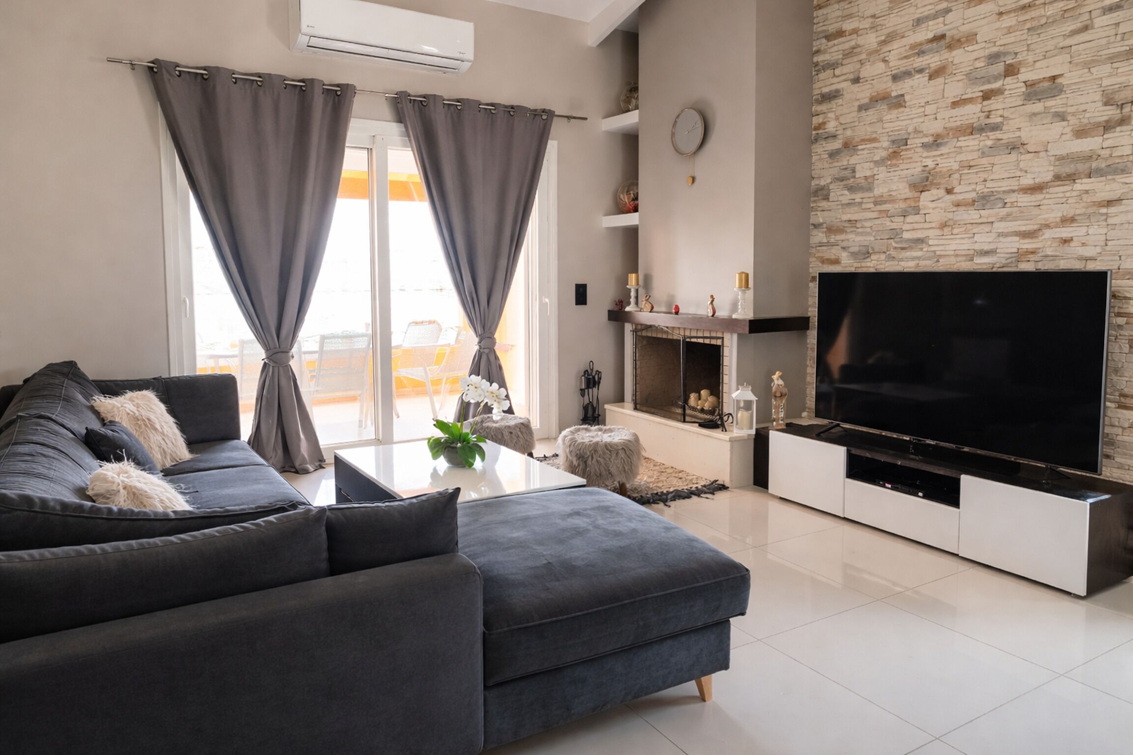 Una sala de estar moderna con un sofá gris, un televisor de pantalla plana, una chimenea y una pared de piedra.