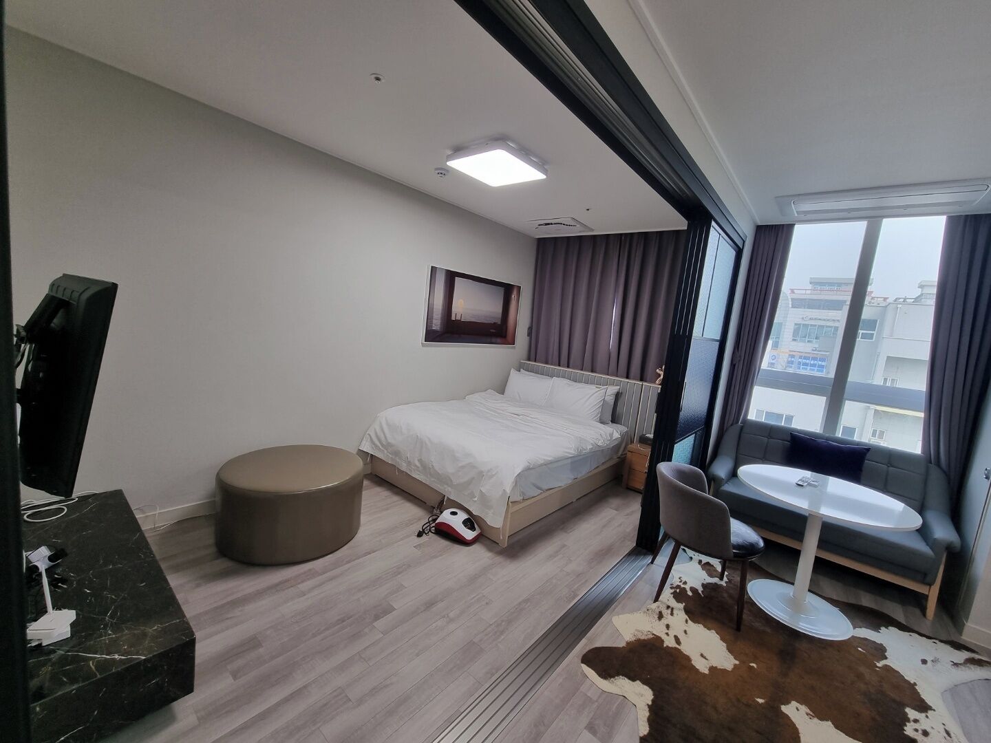 Una habitación de hotel moderna con una cama, un rincón de estar circular, una mesita y un amplio ventanal con cortinas.