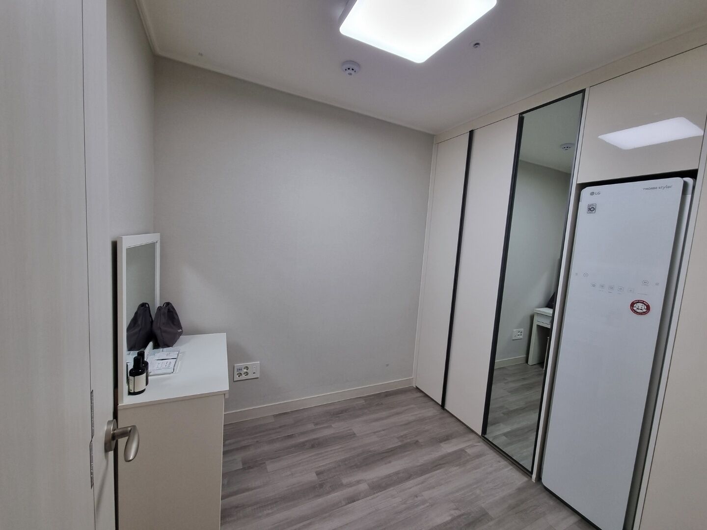 Ein modernes Hotelzimmer mit Holzboden, einem weißen Schrank, einem Spiegel und einer Tür mit Milchglasfenster.