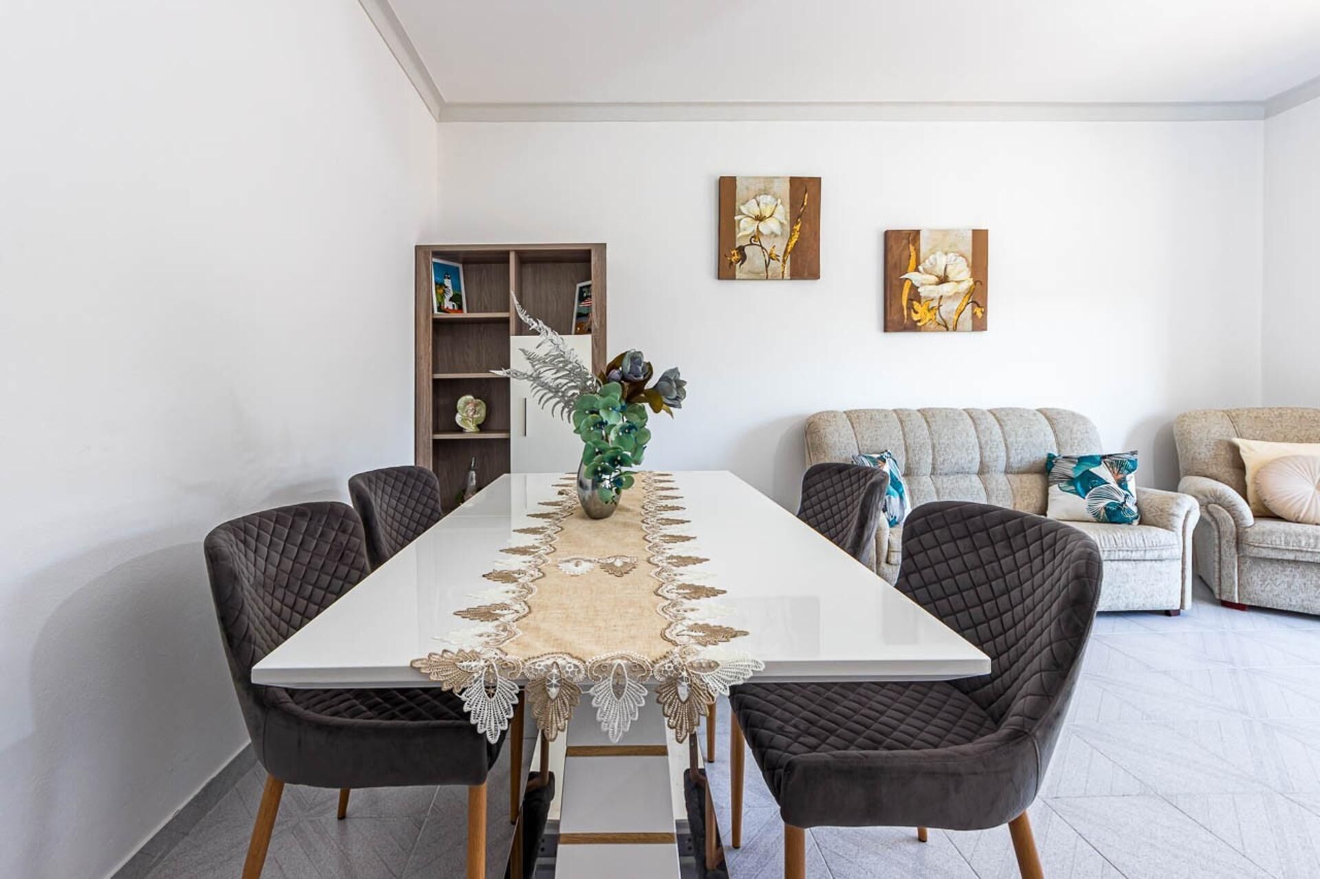 Comedor con una mesa blanca, dos sillas, un mantel decorativo y un jarrón con flores. Hay una estantería con varios objetos y dos cuadros colgados en la pared.