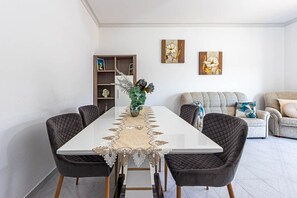 A dining area with a white table, two chairs, a decorative table runner, and a vase with flowers. There is a bookshelf with various items and two wall-mounted paintings.