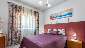 A bedroom with a large bed, floral curtains, a red headboard, a painting, and a nightstand with a lamp. - New Sunset Haven in Gale-2br With Big Terrace (Albufeira)