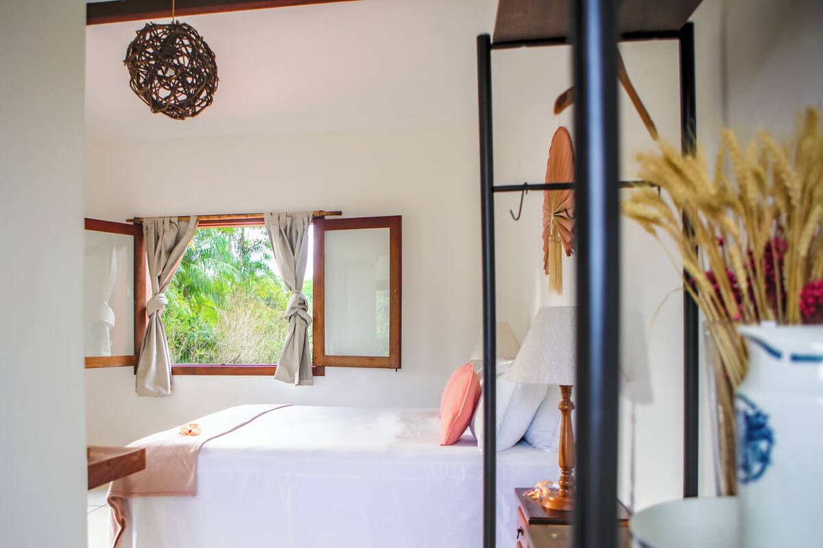 Une chambre à coucher avec un lit, une fenêtre avec des rideaux, un luminaire suspendu et un vase contenant des plantes séchées.