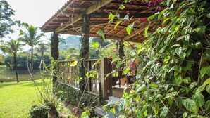 Un pabellón de madera con techo de tejas, rodeado de abundante vegetación y una terraza de madera.