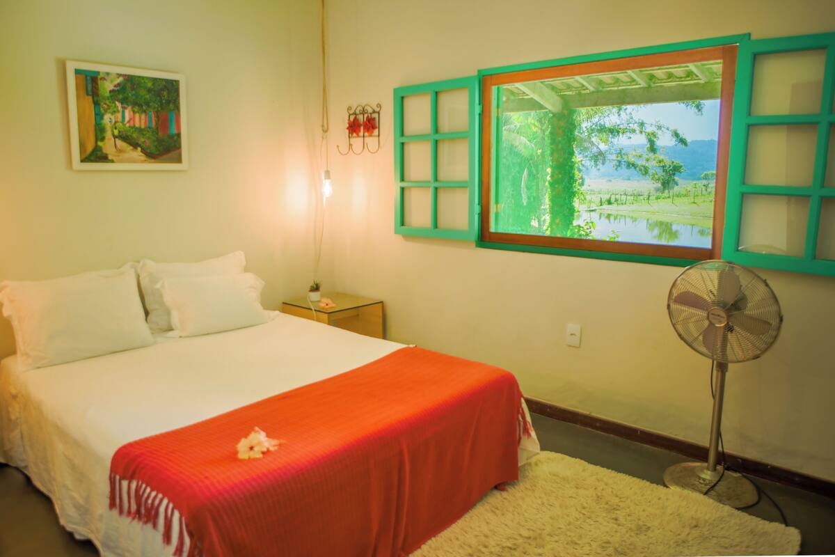 Une chambre à coucher avec un lit, une table de chevet, un ventilateur fixé au mur, une fenêtre donnant sur un paysage verdoyant et un tableau accroché au mur.