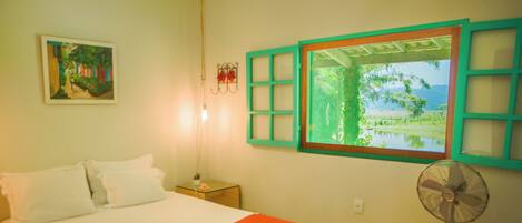A bedroom with a bed, a nightstand, a wall-mounted fan, a window with a view of greenery, and a painting on the wall.