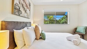A neatly made bed with pillows, a window with a view of greenery, and a painting on the wall. - Tropical Resort Apartment with Pool and Gym Access (Westcourt)