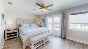 Un dormitorio con cama, mesitas de noche, un banco, ventilador de techo y balcón con vista al océano.