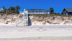 Un hotel frente a la playa con una escalera que lleva a un balcón, un edificio grande con muchas ventanas y un cielo azul despejado.