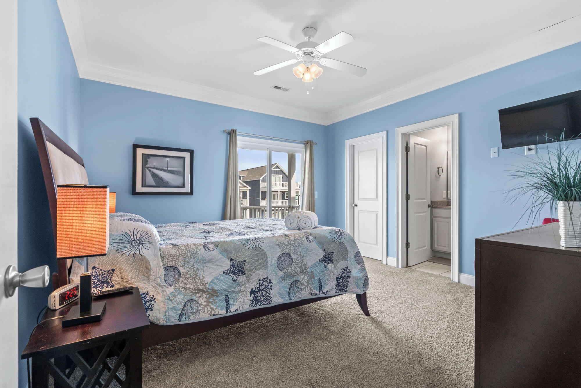 A bedroom with a bed, a nightstand with a lamp, a ceiling fan, a framed picture, and a view of the outside.