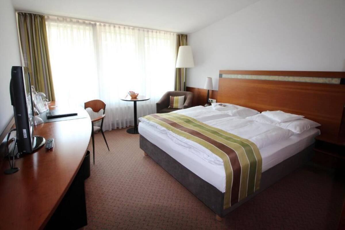 A hotel room with a bed, a desk with a computer, a chair, a small table, and a lamp.