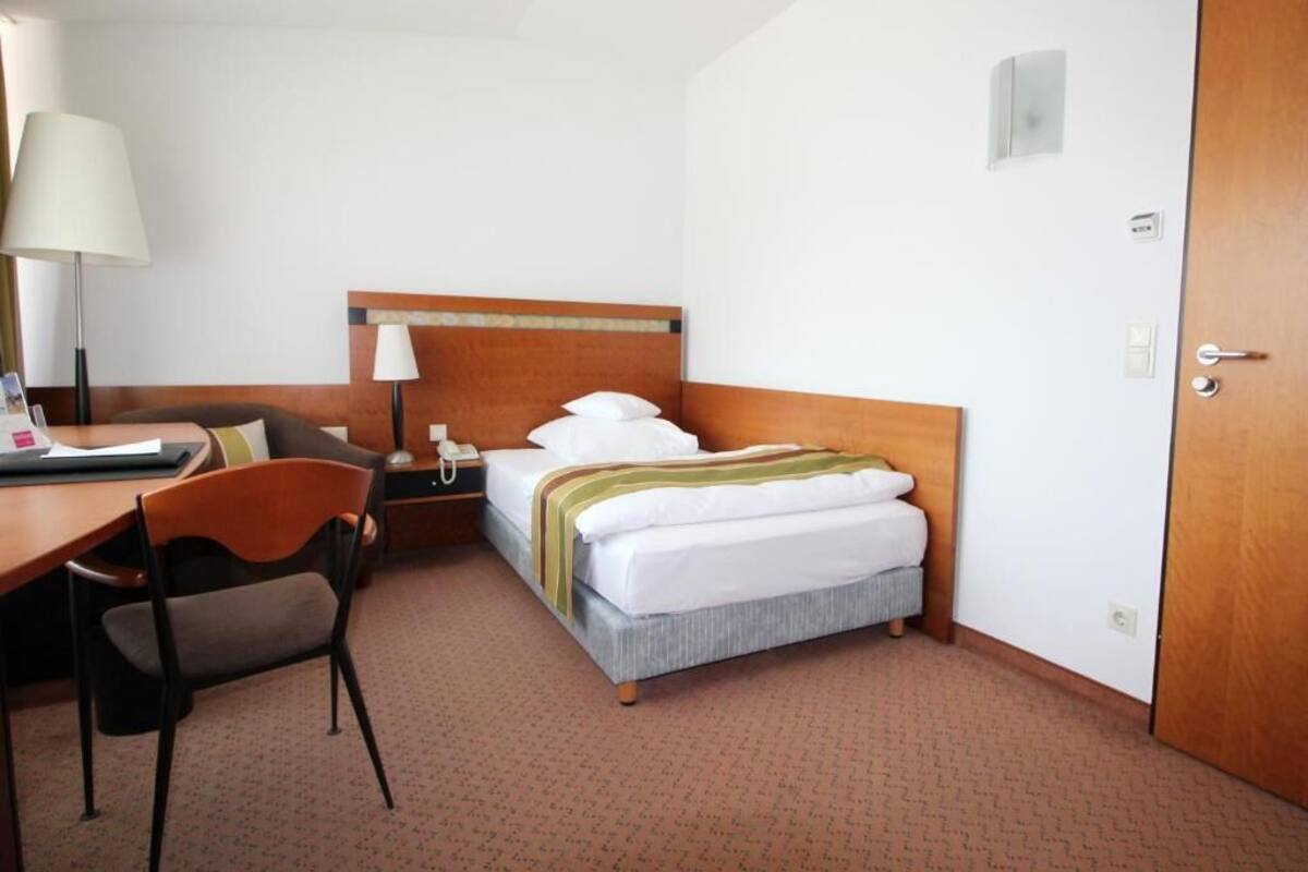 A hotel room with a bed, a desk, a chair, a lamp, and a door.