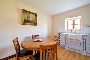 Un coin repas avec une table et des chaises en bois, un tableau au mur et une cuisine avec une fenêtre.